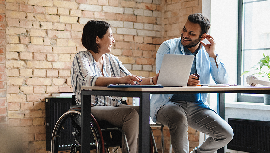 Photo of a woman in a wheelchair and a man are sitting at a desk and talking